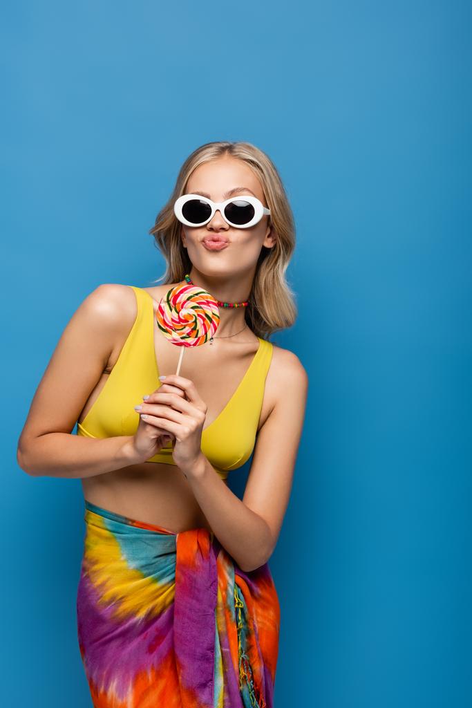 mujer joven en top bikini amarillo y gafas de sol haciendo pucheros labios mientras sostiene dulce piruleta aislada en azul - Foto, Imagen