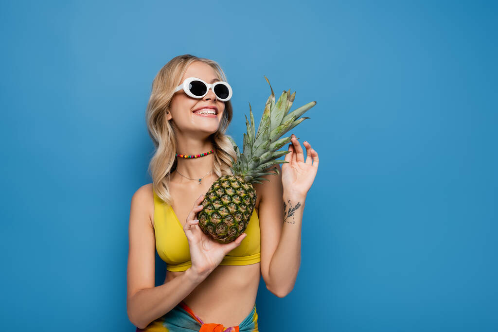 татуйована жінка в сонцезахисних окулярах і бікіні зверху тримає стиглий ананас, посміхаючись ізольовано на синьому
 - Фото, зображення
