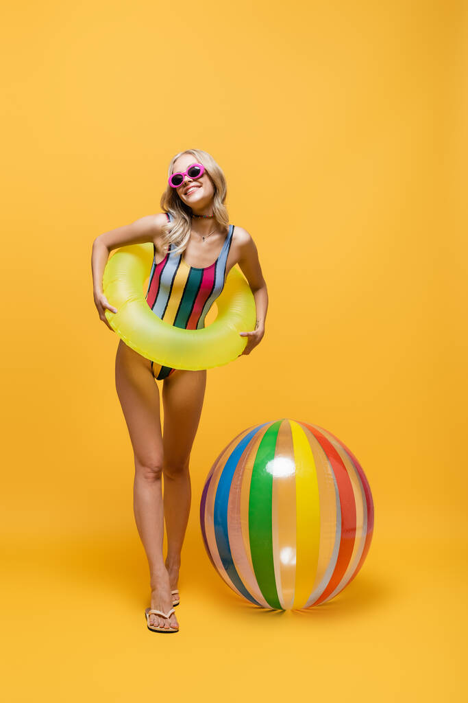 in voller Länge erfreut Frau in Sonnenbrille und Badeanzug mit aufblasbarem Ring in der Nähe von Strandball auf gelb - Foto, Bild