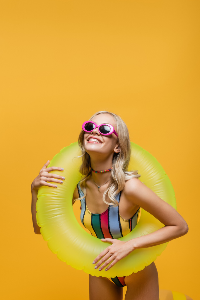 radosna młoda kobieta w okularach przeciwsłonecznych i strojach kąpielowych trzymająca nadmuchiwany pierścień odizolowany na żółto - Zdjęcie, obraz