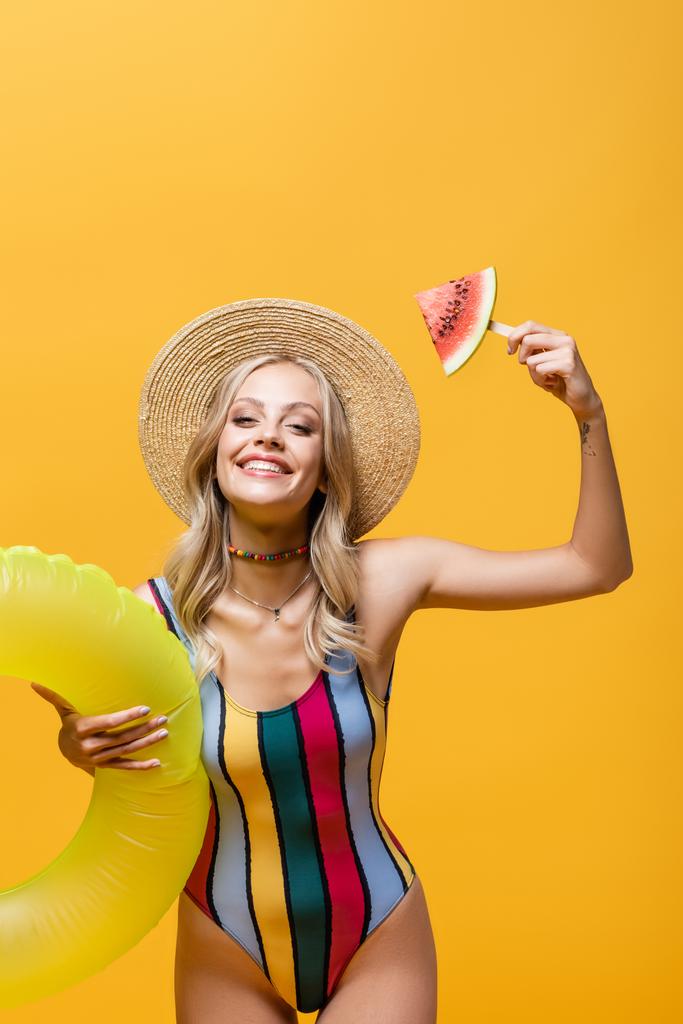 gelukkig vrouw staan met opblaasbare ring en houden lolly stick met watermeloen geïsoleerd op geel  - Foto, afbeelding