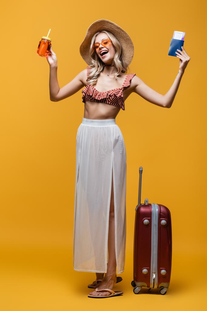 полная длина счастливой женщины в шляпе, стоящей рядом с багажом, держа паспорт и коктейль на желтом  - Фото, изображение