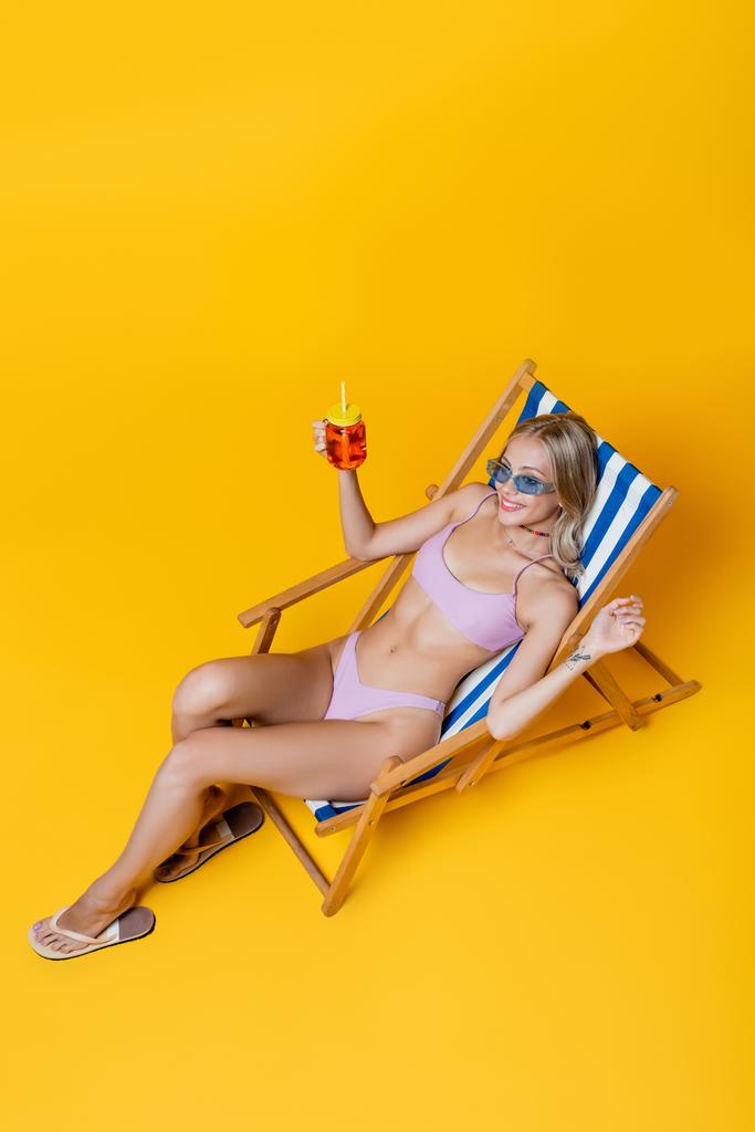 vista de ángulo alto de la joven feliz en traje de baño sentado en la silla de cubierta con cóctel en amarillo  - Foto, Imagen