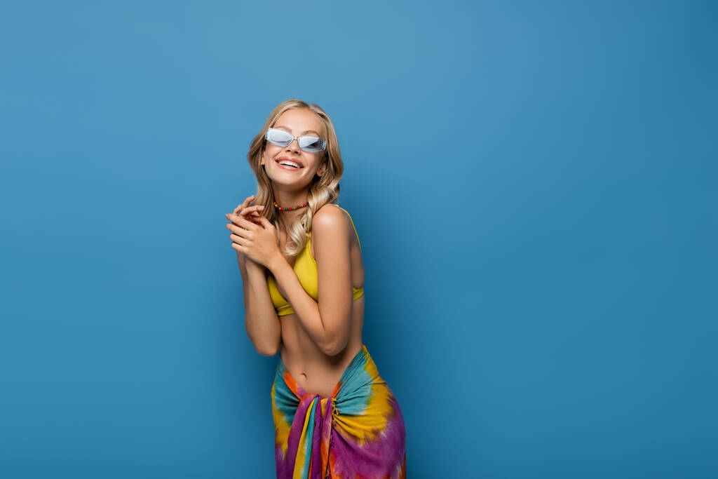 radosna młoda kobieta w okularach przeciwsłonecznych i koralikowym kolorowym naszyjniku uśmiechnięta odizolowana na niebiesko - Zdjęcie, obraz