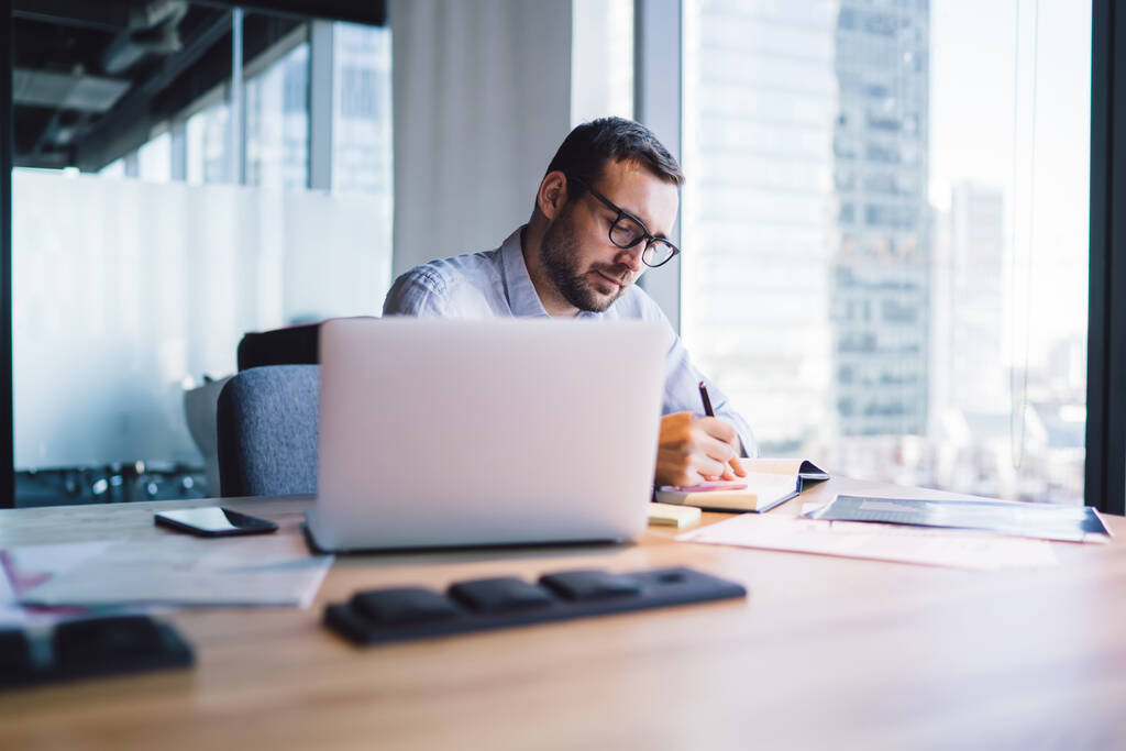 Фокусований інтелектуальний бородатий чоловік в окулярах у синій сорочці робить нотатки в блокноті в офісі, сидячи за столом з ноутбуком
 - Фото, зображення