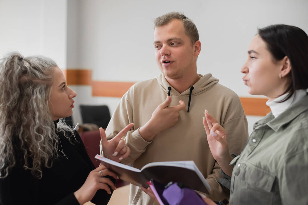 Zwei Mädchen und ein Mann unterhalten sich in Gebärdensprache. Drei gehörlose Studenten unterhalten sich in einem Hörsaal der Universität. - Foto, Bild