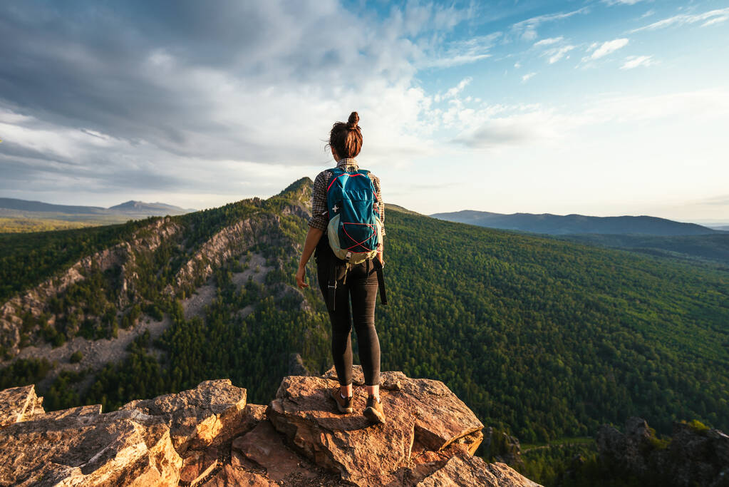 Ein junges Touristenmädchen mit Rucksack genießt den Sonnenuntergang von der Bergspitze aus. Ein Reisender im Hintergrund der Berge. Ein Tourist mit Rucksack steht auf dem Gipfel des Berges - Foto, Bild
