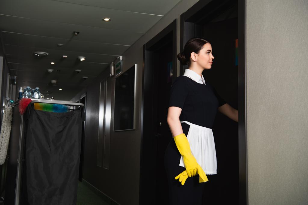 joven sonriente criada golpeando puerta en pasillo de hotel  - Foto, Imagen