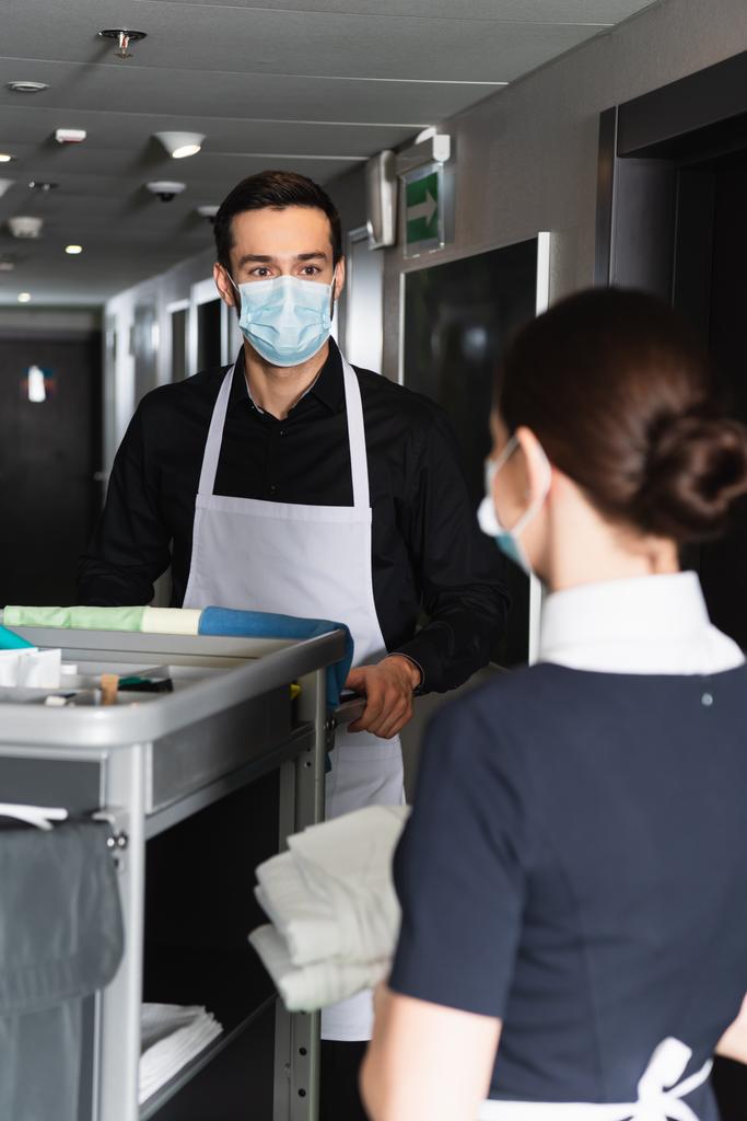 governanta em máscara médica olhando para empregada doméstica borrada perto do carrinho de limpeza  - Foto, Imagem
