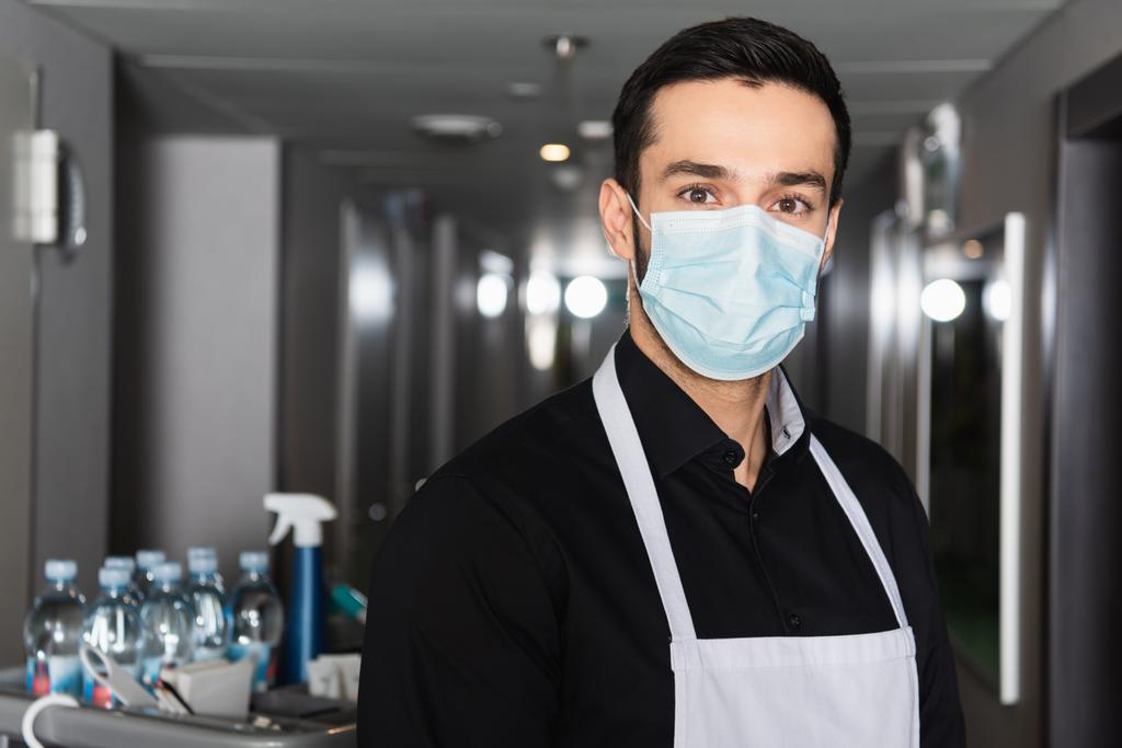 housekeeper in apron and medical mask looking at camera in corridor of hotel - Photo, Image