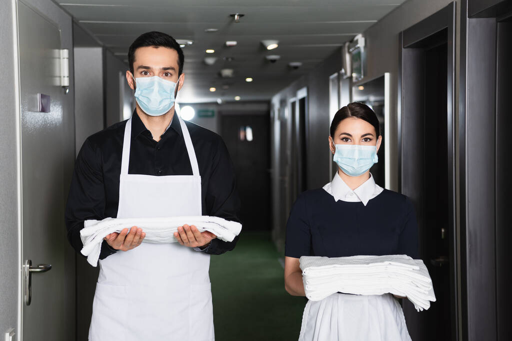 Otel koridorunda temiz çarşaf tutan üniformalı ve tıbbi maskeli hizmetçiler.  - Fotoğraf, Görsel