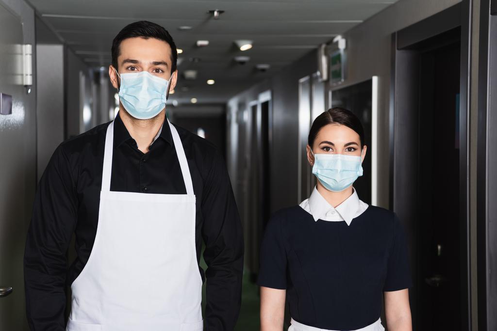 young housekeepers in uniform and medical masks looking at camera in corridor of hotel  - Photo, Image