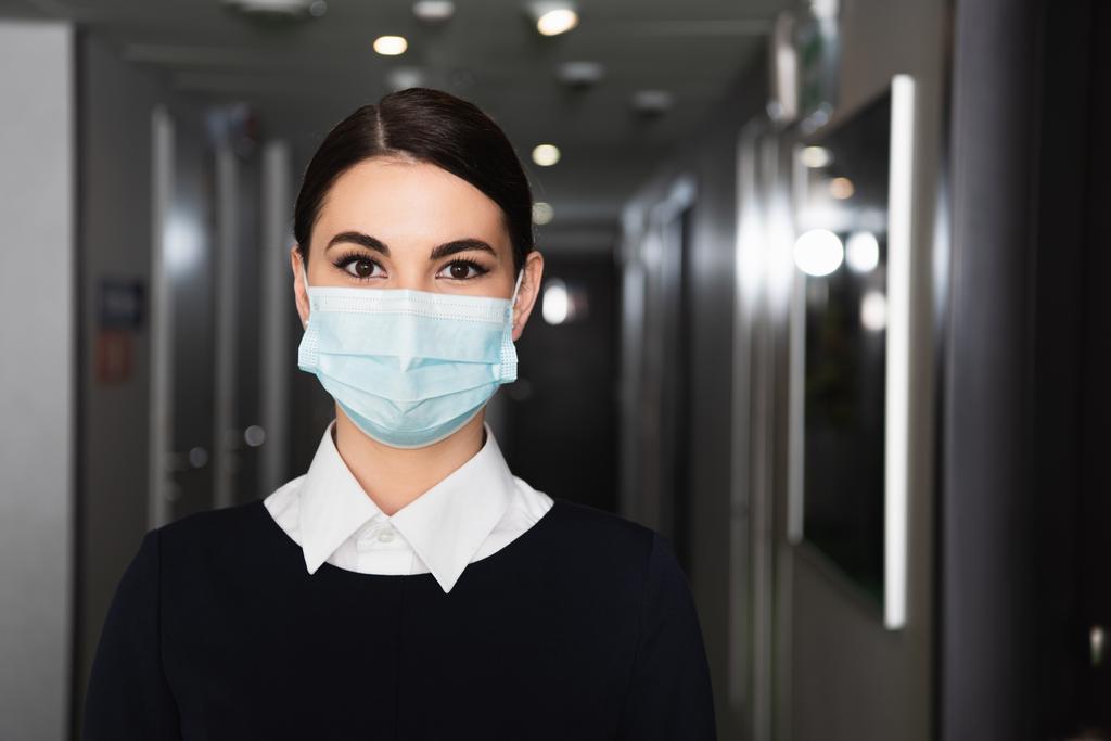 junges Dienstmädchen in medizinischer Maske und Uniform blickt auf Hotelflur in die Kamera  - Foto, Bild