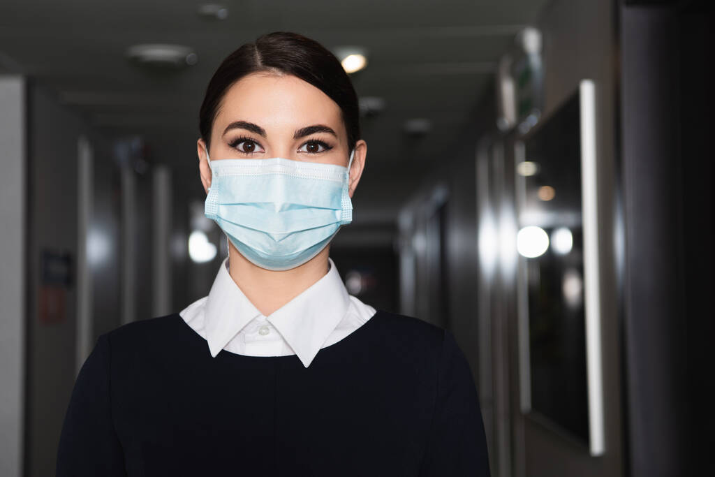 morena criada en máscara médica y uniforme mirando cámara en pasillo de hotel  - Foto, Imagen