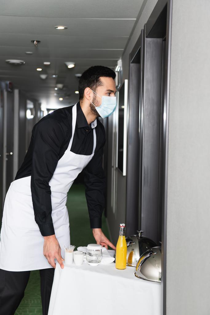 kelner w masce medycznej i fartuchu dostarczający tacę z kloszem i napojami w hotelu  - Zdjęcie, obraz