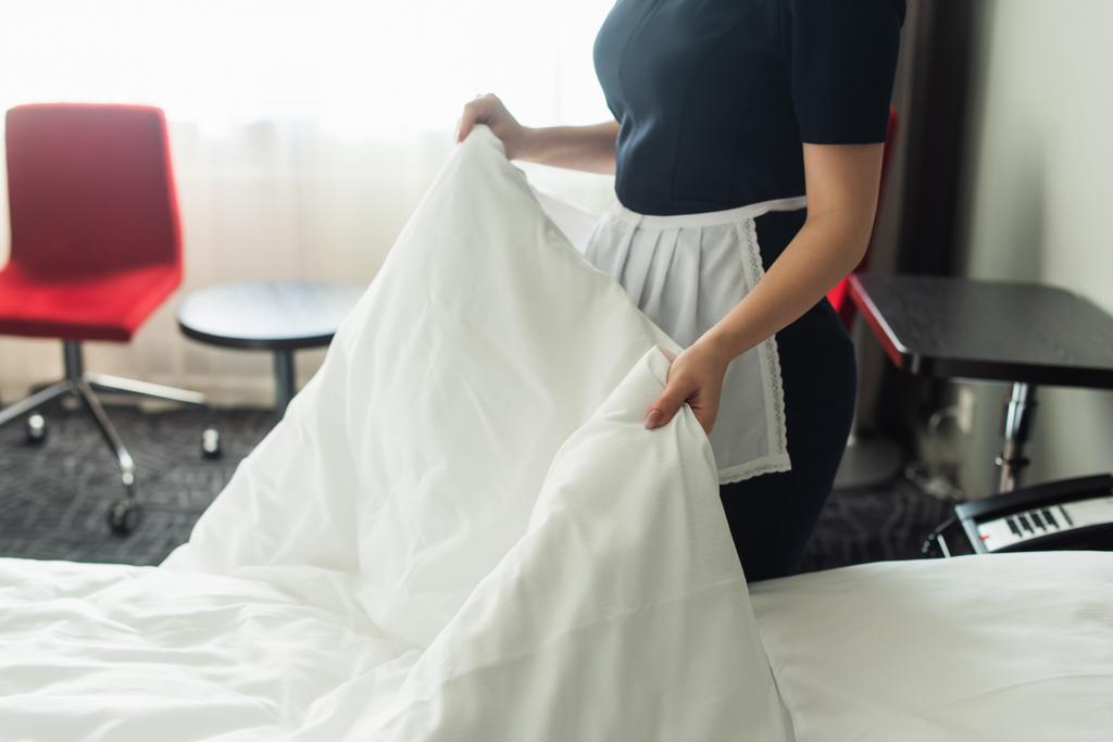 vista recortada de la joven camarera en uniforme cambiante ropa de cama en la habitación de hotel  - Foto, Imagen