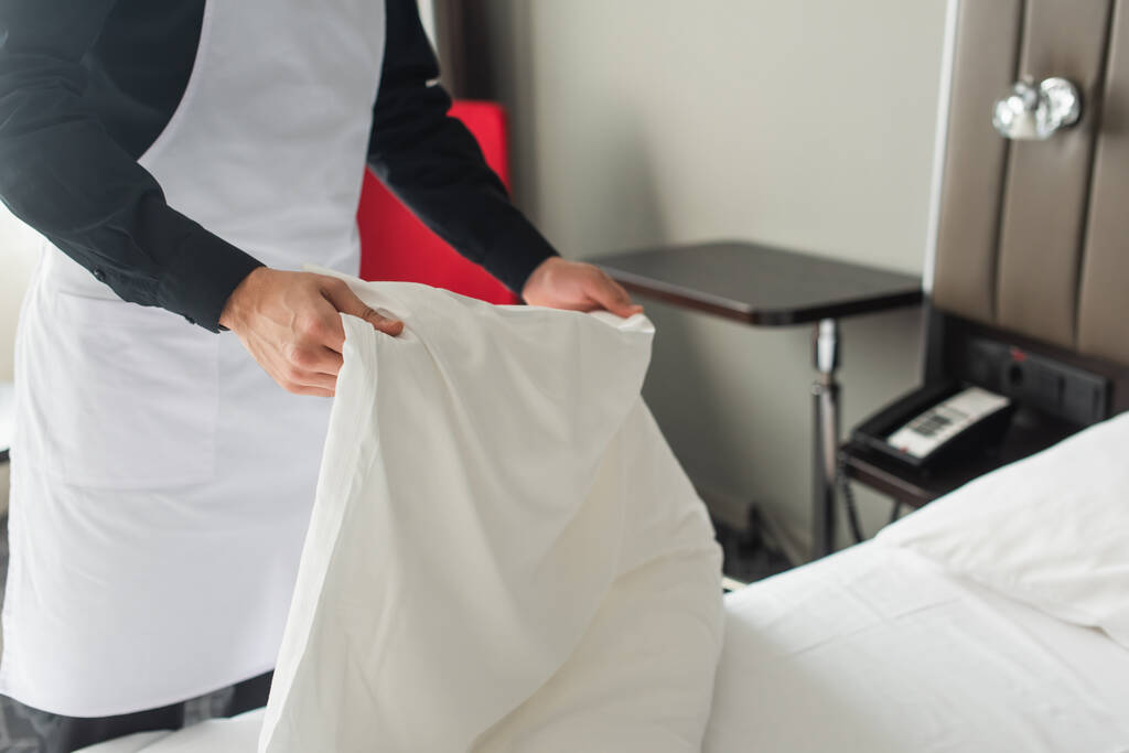 partial view of housekeeper in apron changing bedding in hotel room  - Photo, Image
