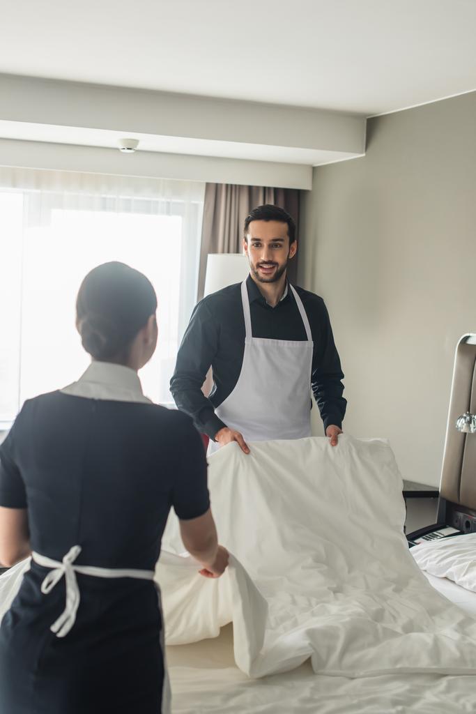 χαμογελαστή και γενειοφόρος οικονόμος αλλαγή κρεβάτι με θολή υπηρέτρια στο δωμάτιο του ξενοδοχείου  - Φωτογραφία, εικόνα