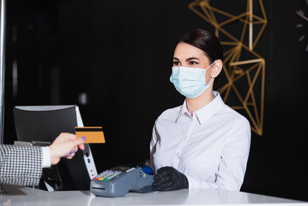 ρεσεψιονίστ σε γάντια λάτεξ και ιατρική μάσκα κρατώντας τερματικό πληρωμής κοντά επισκέπτη με πιστωτική κάρτα  - Φωτογραφία, εικόνα
