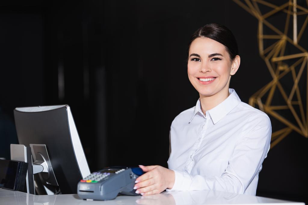 feliz recepcionista sonriendo cerca de lector de tarjetas de crédito en el mostrador  - Foto, Imagen