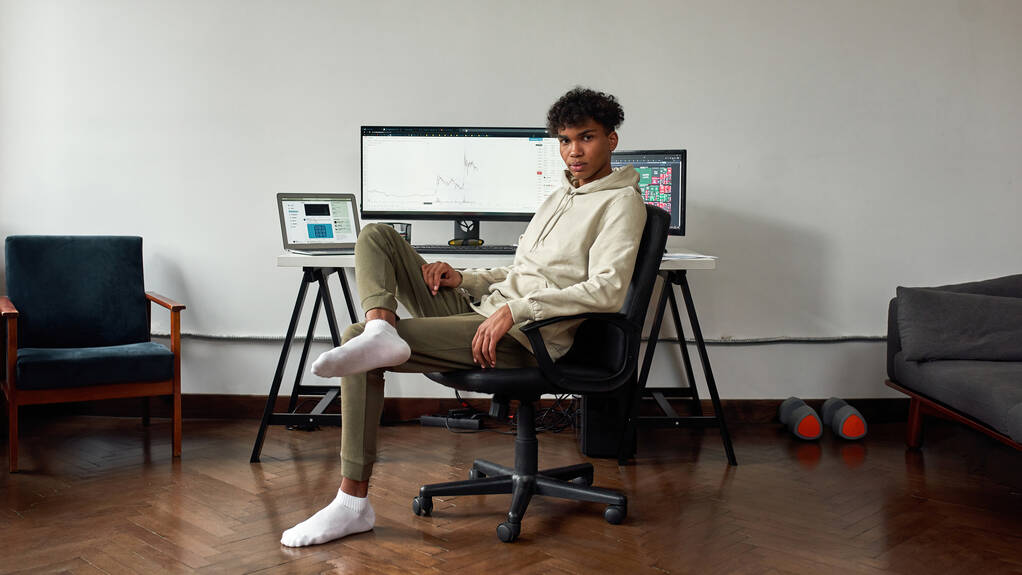 Full length shot of young mixed race man trading online χρησιμοποιώντας φορητό υπολογιστή και υπολογιστή, που κάθεται στο σπίτι στο σύγχρονο γραφείο σαλονιού - Φωτογραφία, εικόνα