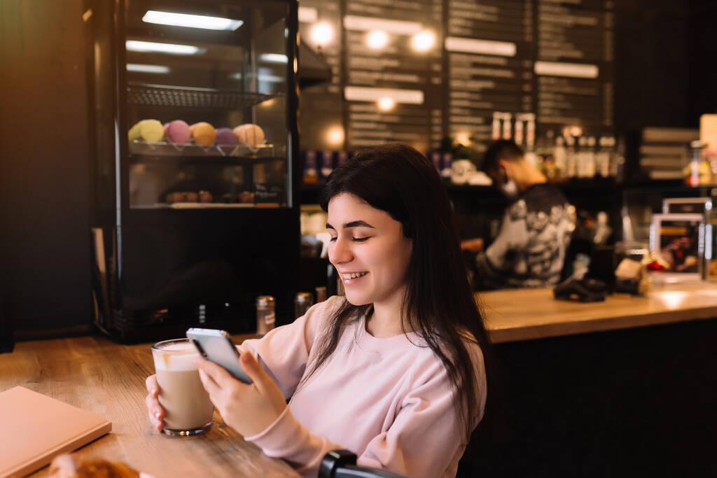 A lány mosolyog, kávét iszik a kávézóban és telefonokat olvas. Homályos háttér. Kiváló minőségű fénykép - Fotó, kép