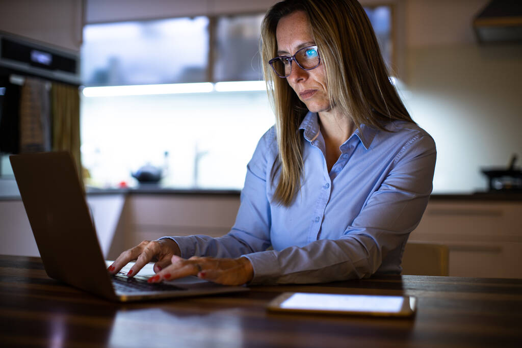 Mooie vrouw van middelbare leeftijd die laat op de dag werkt op een laptop thuis, een bedrijf runt vanuit huis, op afstand werkt - gefrustreerd raakt - Foto, afbeelding
