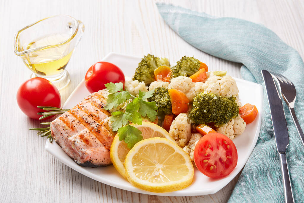 Gegrilltes Lachsfilet garniert mit Gemüse, Zitrone, Tomaten, Kräutern, die auf einem Teller auf hölzernem Hintergrund liegen. Gesunde Ernährung. Keto-Diät. - Foto, Bild