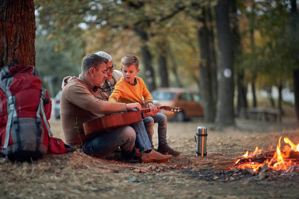 An elderly couple and grandson playing guitar in the forest around a campfire on a beautiful autumn dusk - Photo, Image