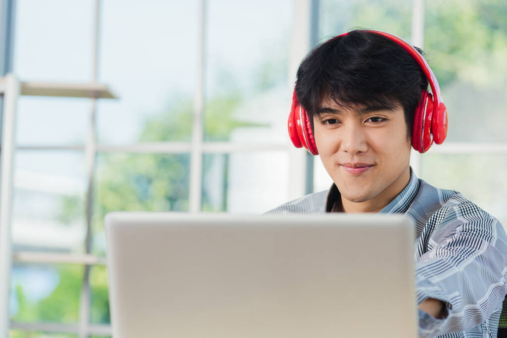 Азіатський молодий бізнесмен щасливий з червоними навушниками, які він сидить на робочому місці, шукаючи монітор ноутбука, впевнений, що красивий чоловік спосіб життя, посміхається він слухає музику - Фото, зображення