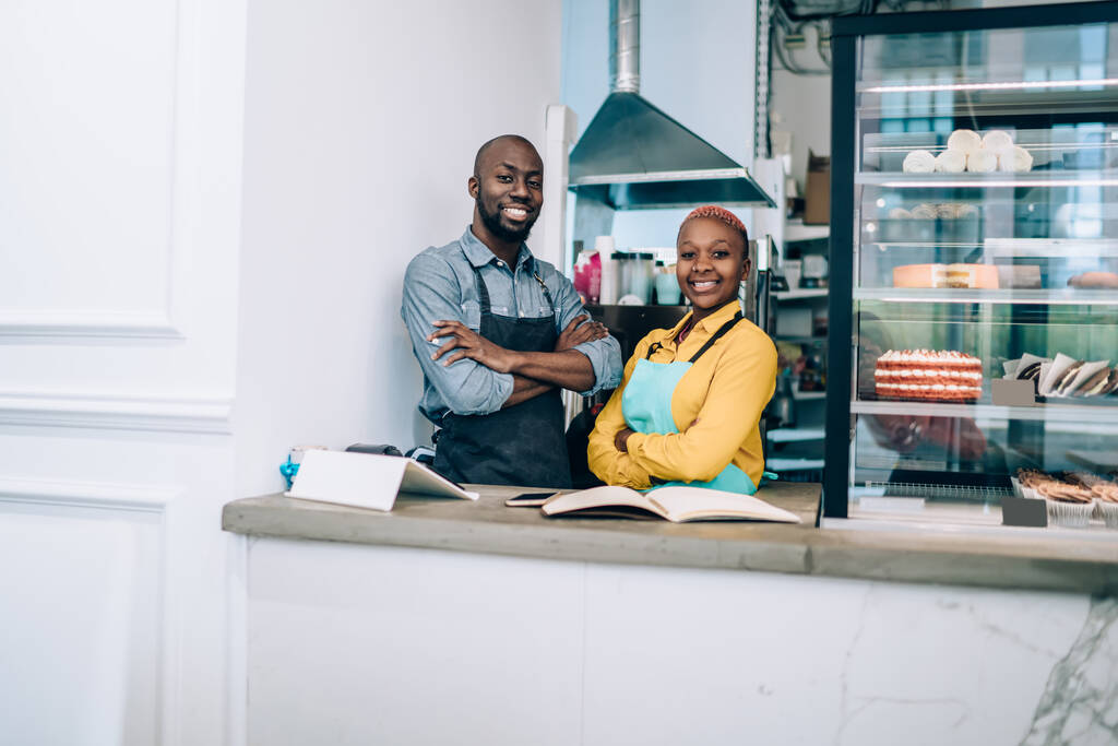 Улыбающиеся афроамериканские повара в желтых и серых рубашках и синих и черных фартуках, скрещивающие руки и смотрящие в камеру со смехом на кухне  - Фото, изображение