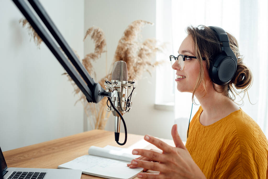Woman podcaster streaming and broadcasting her podcast from homemade studio. Female recording podcast from home and talking into a microphone. - Photo, Image