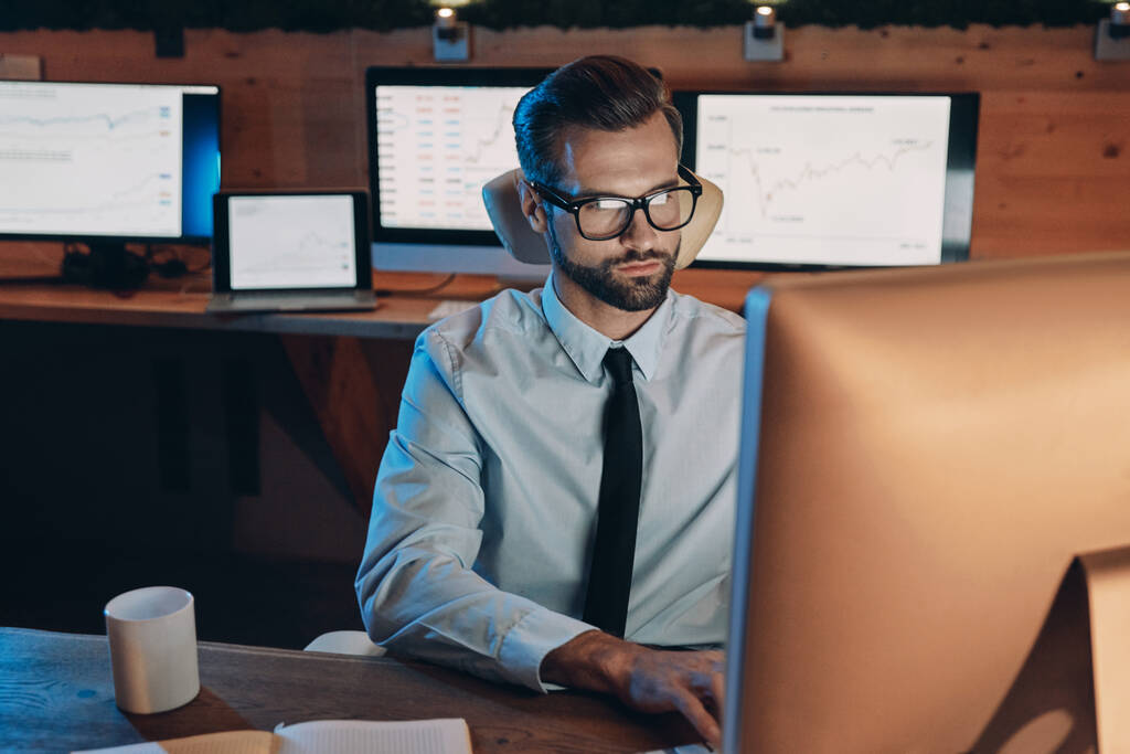 Уверенный молодой человек, работающий за компьютером, оставаясь допоздна в офисе - Фото, изображение