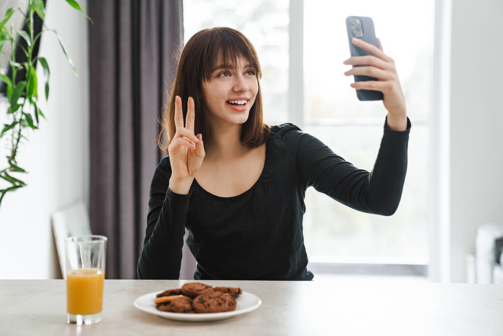 Mujer blanca joven feliz mirando el teléfono móvil mientras está sentado en la mesa de la cocina en casa tomando una selfie - Foto, imagen