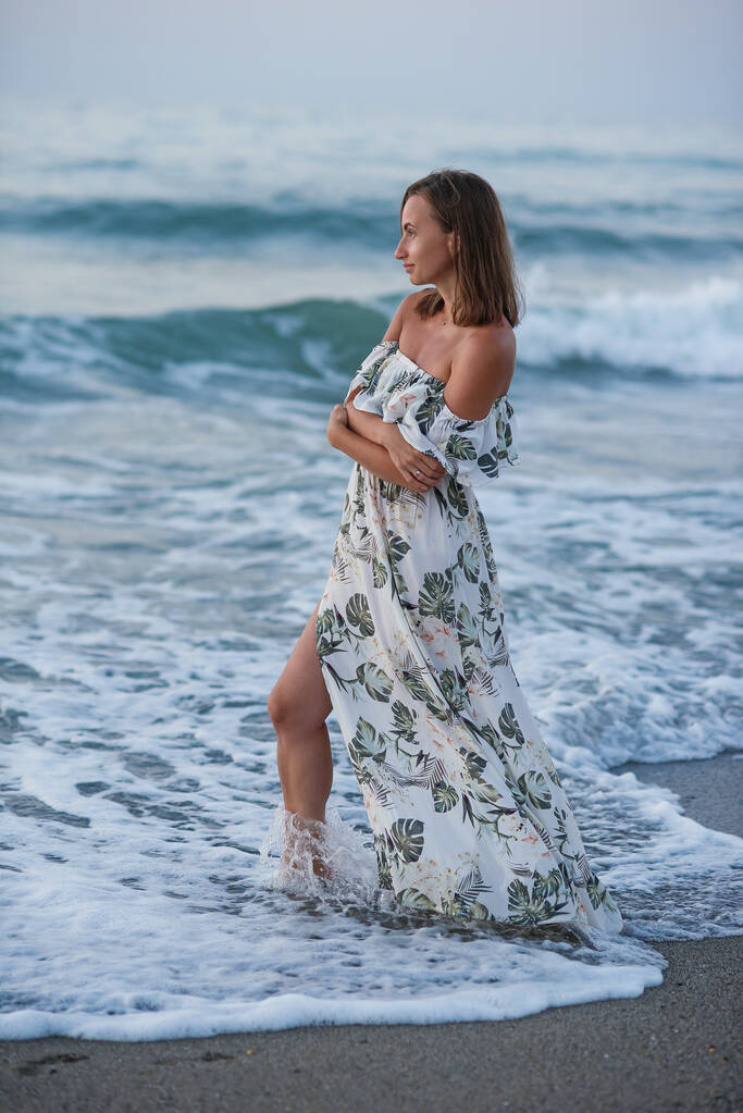 Joven mujer bronceada hermosa usando un vestido elegante posando de pie en la playa cerca de las olas y mirando hacia el mar. - Foto, Imagen