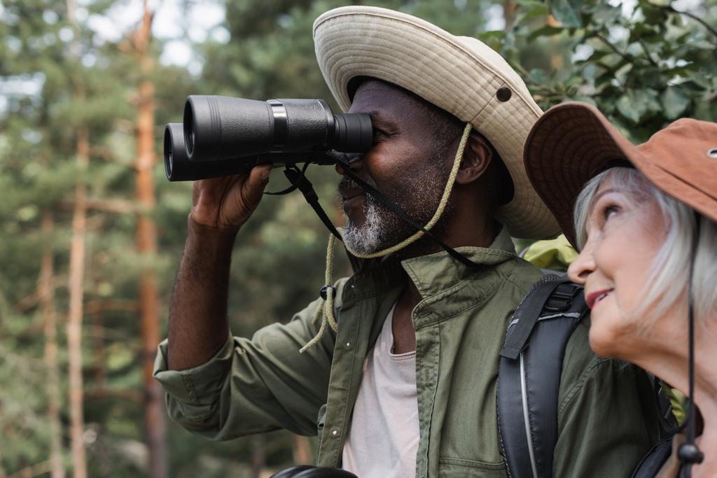Afrikanischer Wanderer sucht mit Fernglas in der Nähe seiner lächelnden Frau  - Foto, Bild