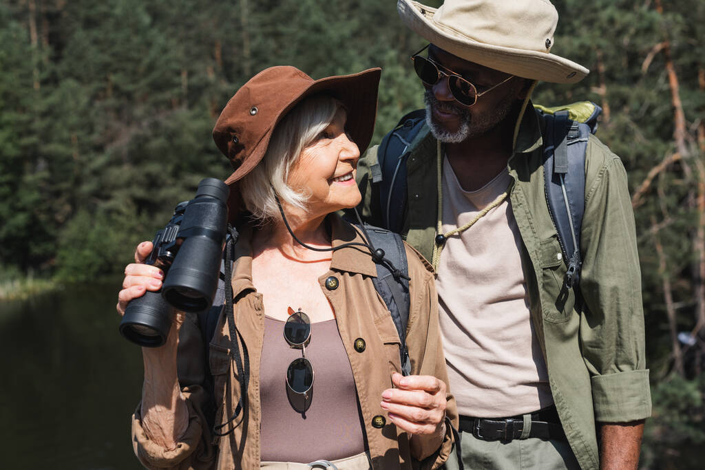 Feliz viajero senior sosteniendo prismáticos cerca de esposo afroamericano en bosque  - Foto, imagen