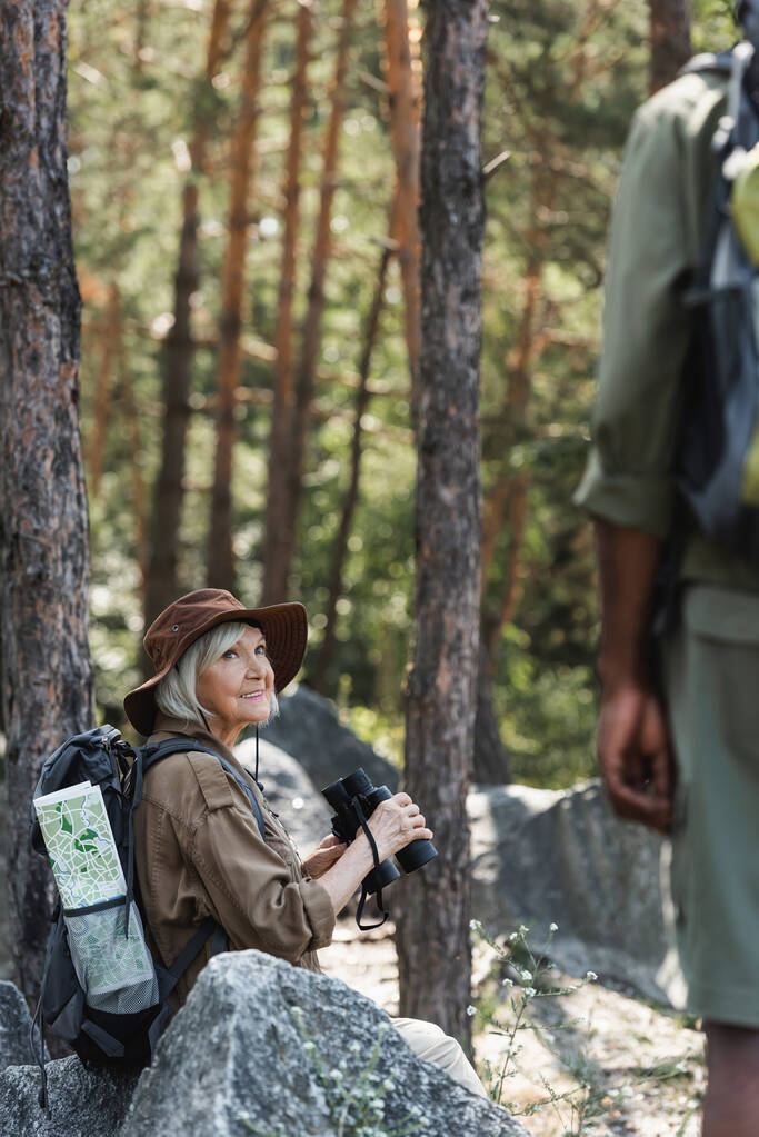 Mujer mayor sonriente sosteniendo prismáticos cerca del borroso esposo afroamericano en el bosque  - Foto, Imagen