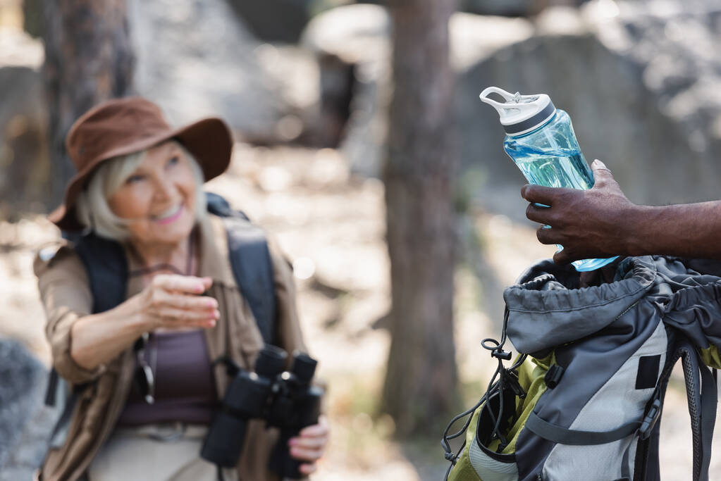 Αφρο-αμερικανός ταξιδιώτης κρατώντας ένα μπουκάλι νερό κοντά σε θολή σύζυγο στο δάσος  - Φωτογραφία, εικόνα