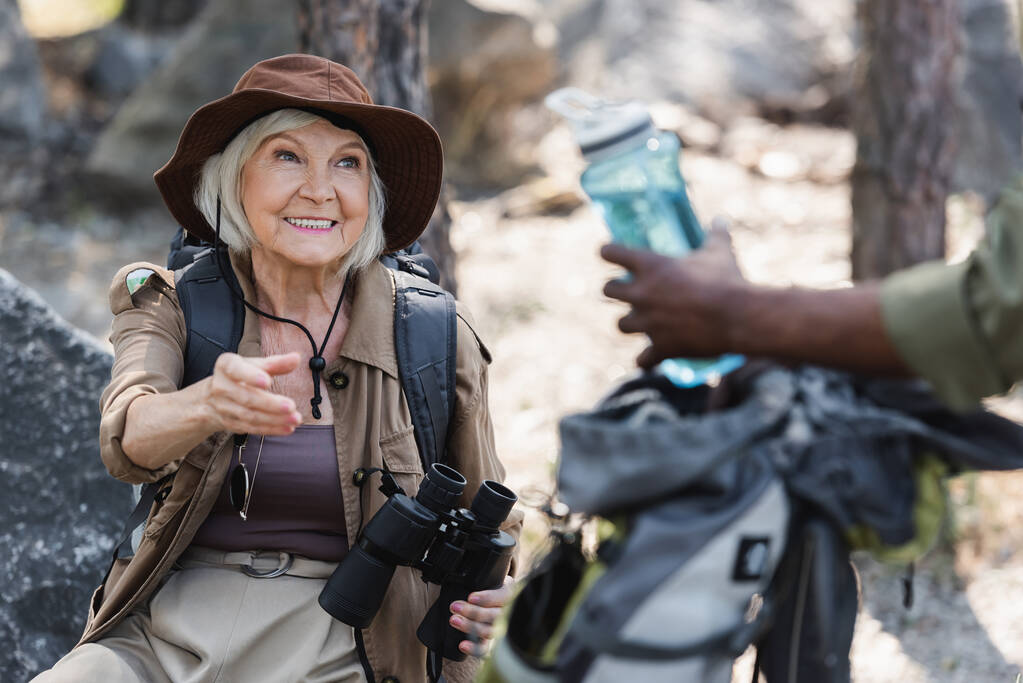 Turista anziano con binocolo mano di outreaching vicino al marito afroamericano con bottiglia d'acqua nella foresta  - Foto, immagini