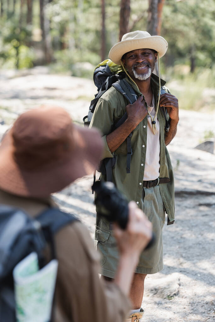 Vrolijke Afrikaanse Amerikaanse toerist met rugzak op zoek naar wazige vrouw in het bos  - Foto, afbeelding