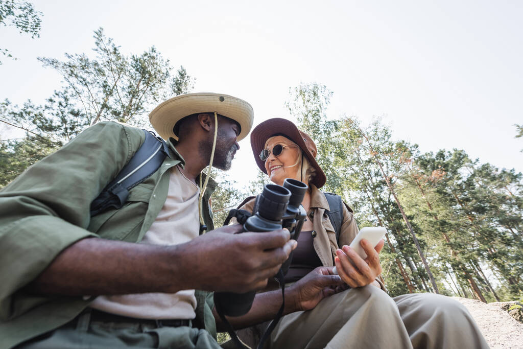 Низький кут зору позитивних багатоетнічних мандрівників, що тримають смартфон і бінокль в лісі
  - Фото, зображення