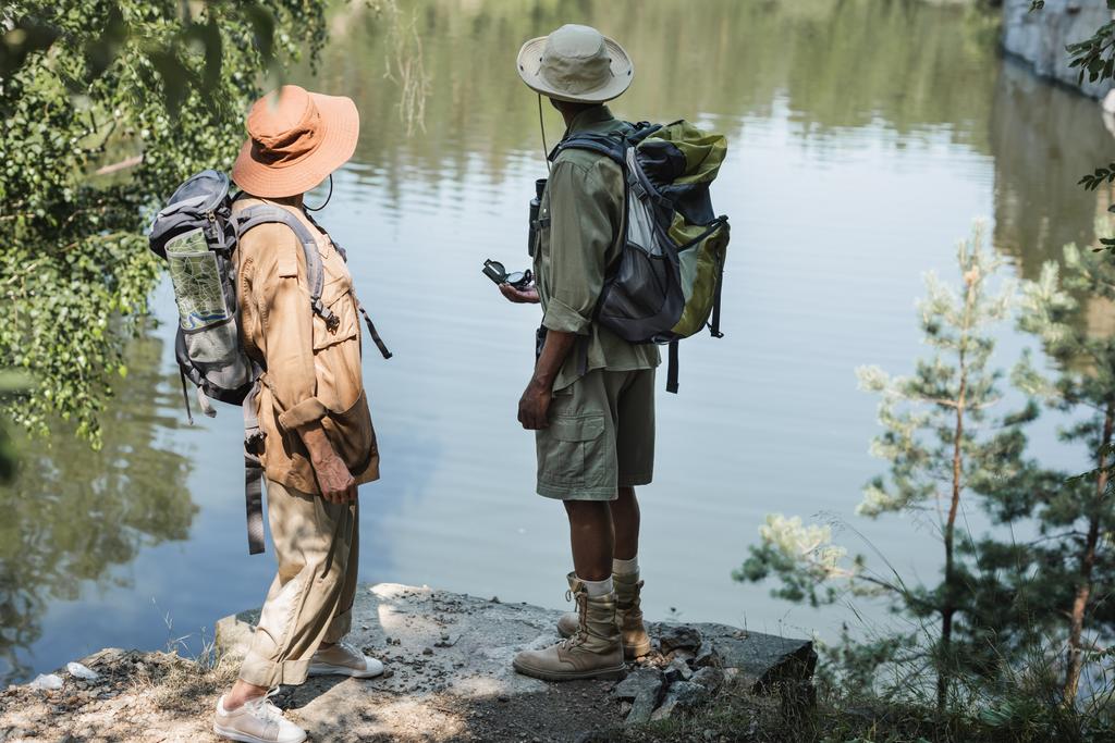 Senior interracial couple with backpacks and compass standing near lake  - Photo, Image