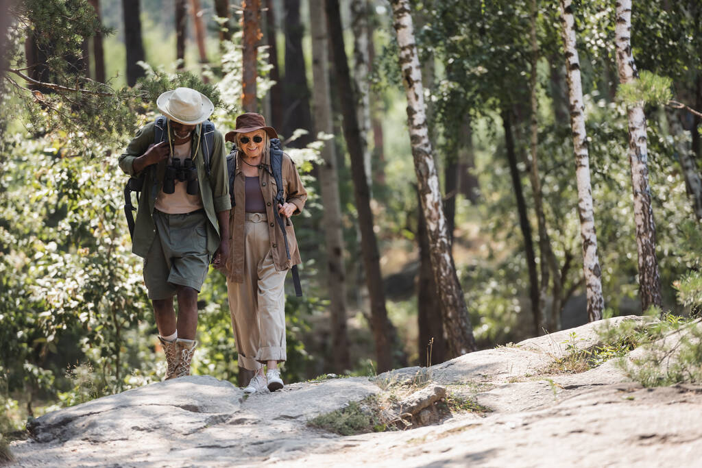 Uśmiechnięta starsza kobieta z plecakiem spacerująca w pobliżu afrykańskiego męża w lesie  - Zdjęcie, obraz
