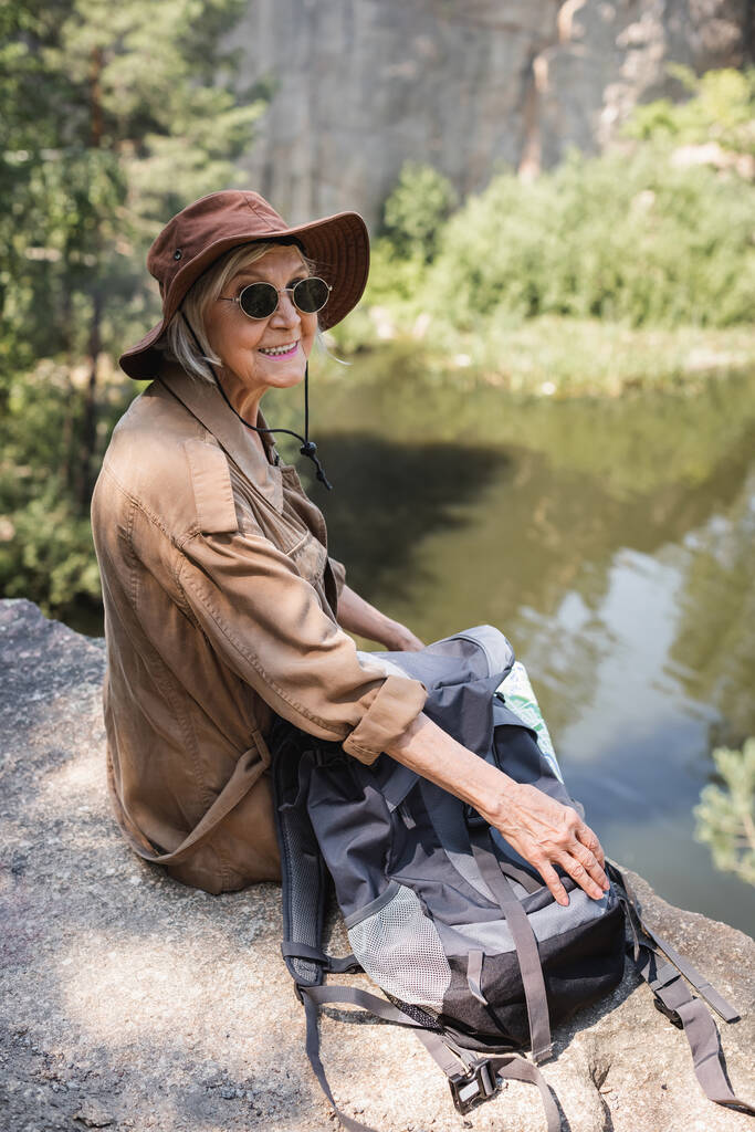 Senior Tourist mit Sonnenbrille sitzt neben Rucksack auf Klippe  - Foto, Bild