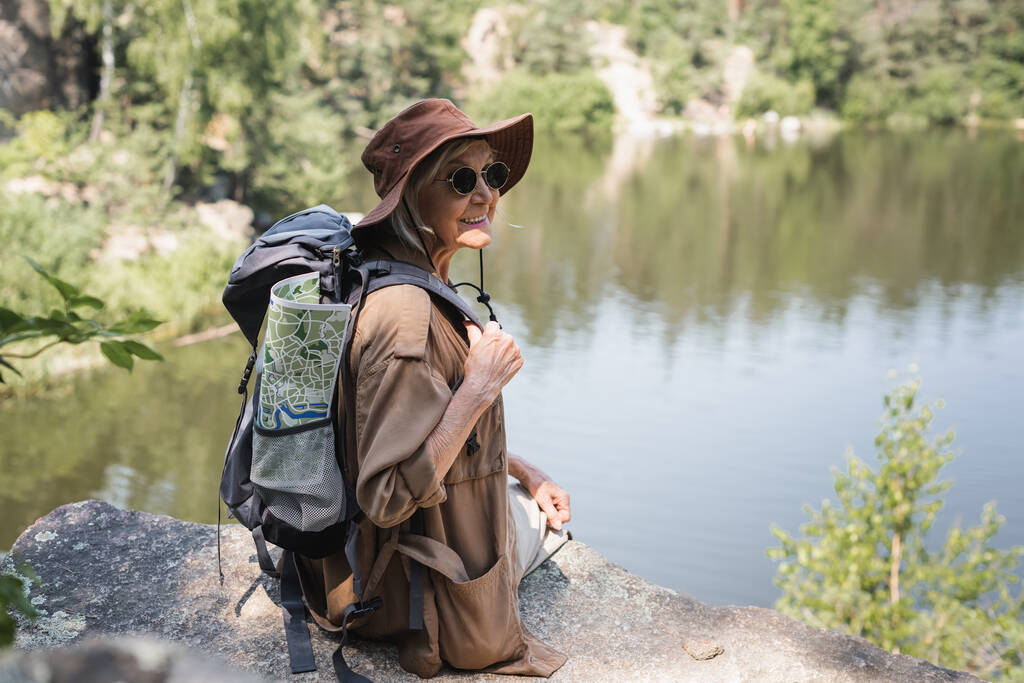 Positiver Wanderer mit Rucksack und Karte sitzt auf Felsen am See  - Foto, Bild
