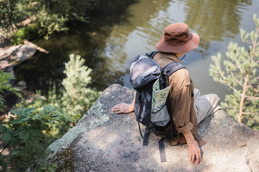 Mulher sênior com mochila e mapa sentado na rocha perto do lago  - Foto, Imagem