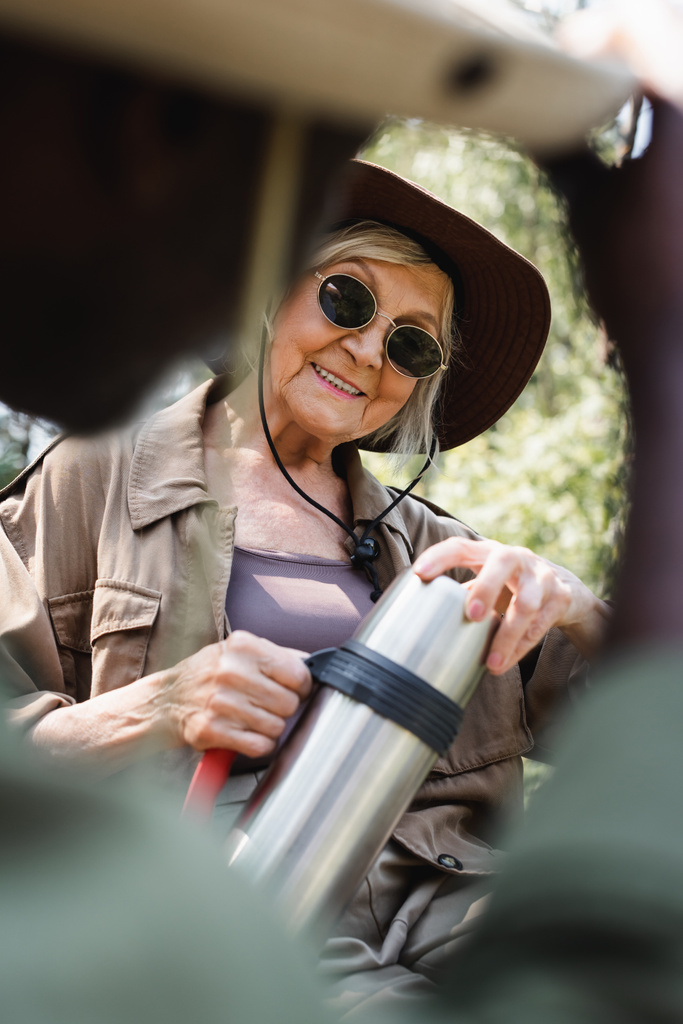 Pozitivní senior turista držení termosku v blízkosti rozmazané africký americký manžel  - Fotografie, Obrázek