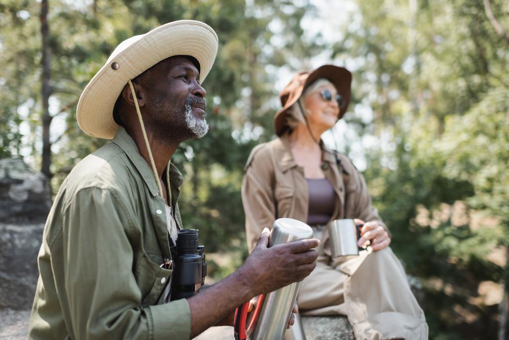 Lächelnder afrikanisch-amerikanischer Tourist mit Thermoskanne und Fernglas neben verschwommener Frau im Wald  - Foto, Bild