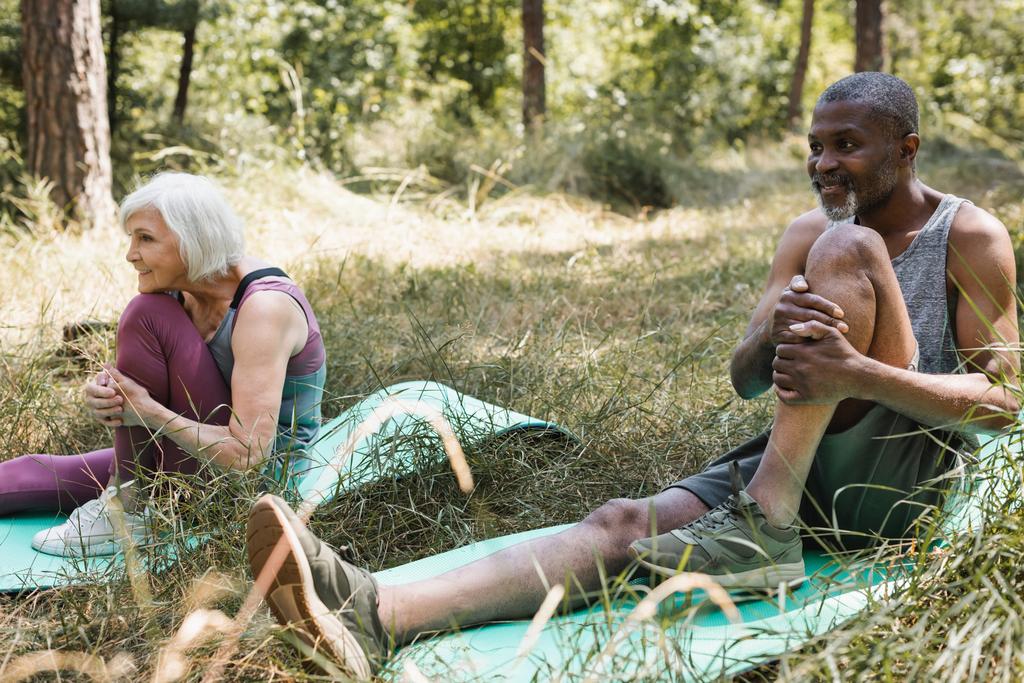 Улыбающийся африканский американец растягивается на тренажерном коврике рядом с женой в лесу  - Фото, изображение
